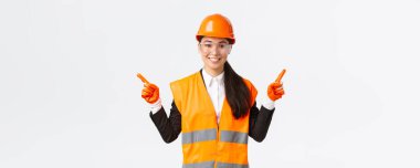 Gülümseyen Asyalı kadın müteahhit, güvenlik kasklı mühendis, koruma gözlüğü yana bakan, sol ve sağ projeyi gösteren, inşaat projelerini tanıtan, beyaz arka plan