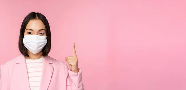 Close up retrato de mulher de negócios asiática em máscara facial médica e terno, apontando o dedo para cima, mostrando propaganda, banner superior, de pé sobre fundo rosa — Fotografia de Stock