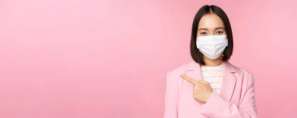 Tıbbi yüz maskesi ve takım elbiseli Asyalı iş kadını portresi, sol parmağı göstererek reklam, şirket afişi, stüdyo pembe arka planı — Stok fotoğraf