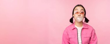 Havalı Asyalı kız sakız şişiriyor, sakız çiğniyor, güneş gözlüğü takıyor, pembe arka planda poz veriyor.