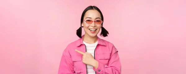 Πορτρέτο του χαμογελαστού κορίτσι της Ασίας σε κομψό ντύσιμο, γυαλιά ηλίου, δείχνοντας το δάχτυλο αριστερά, δείχνει διαφήμιση, πανό, στέκεται πάνω από ροζ φόντο — Φωτογραφία Αρχείου