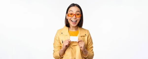 Retrato de bela menina asiática moderna em óculos de sol, sorrindo feliz, mostrando cartão de crédito, de pé sobre fundo branco — Fotografia de Stock