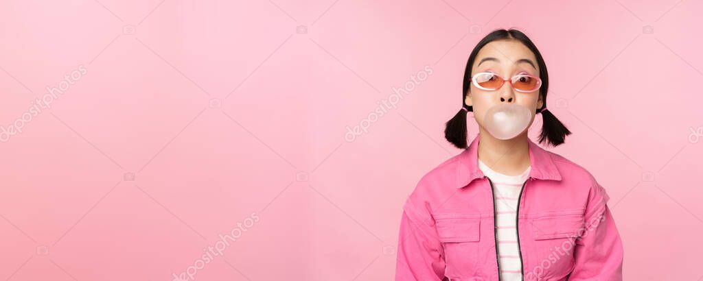 Stylish asian girl blowing bubblegum bubble, chewing gum, wearing sunglasses, posing against pink background. Copy space