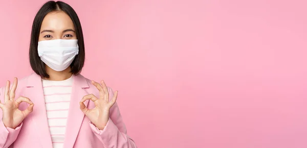 Asiatisk affärskvinna i kostym och medicinsk ansiktsmask, visar okej tecken, rekommenderar att använda skyddsutrustning på kontoret under covid-19 pandemi, rosa bakgrund — Stockfoto