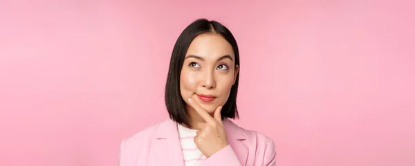 Close up portrait of young asian businesswoman thinking, smiling thoughtful and looking at upper left corner, standing over pink background — Stock Photo, Image