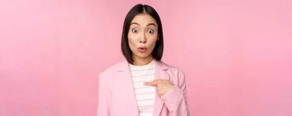 Retrato de mulher de negócios asiática reage surpreso, aponta para si mesma com descrença no rosto, posando em terno contra fundo rosa — Fotografia de Stock