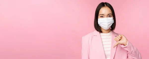 Mulher de negócios coreana em máscara facial médica e terno, mostra polegares para baixo, desgosto ou desaprovar gesto, de pé sobre fundo rosa — Fotografia de Stock