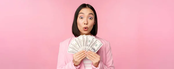 Finance, microcredit and people concept. Happy smiling asian businesswoman showing dollars money, standing in suit against pink background — Stock Photo, Image