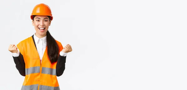 Vincitore di successo asiatico direttore di costruzione femminile, ingegnere a vista edificio indossando casco di sicurezza, alzando le mani nella gioia, pompa pugno dal raggiungimento e la vittoria, sfondo bianco — Foto Stock