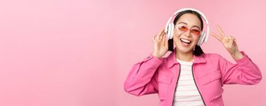 Dans eden Asyalı kız kulaklıkla müzik dinliyor, pembe arka planda poz veriyor.