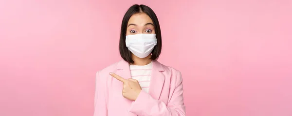 Retrato de mulher de negócios asiática em máscara facial médica e terno, apontando o dedo esquerdo, mostrando propaganda, banner da empresa, estúdio rosa fundo — Fotografia de Stock