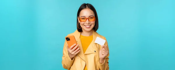 Handla på nätet. Snygg ung asiatisk kvinna i solglasögon, visar kreditkort och använda smartphone, betala på internet, göra inköp, står över blå bakgrund — Stockfoto
