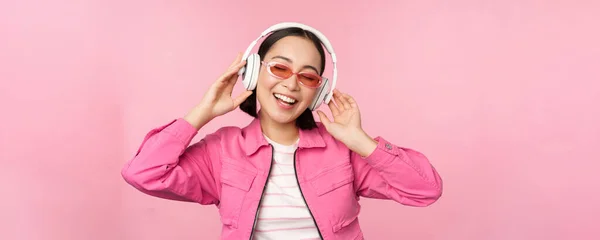 Stilvolles asiatisches Mädchen, das mit Kopfhörern Musik hört und vor rosa Hintergrund posiert — Stockfoto