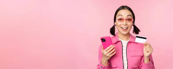 Compras en línea. Sonriendo chica asiática shopper, sosteniendo teléfono inteligente y tarjeta de crédito, pagando en aplicación móvil, de pie sobre fondo rosa — Foto de Stock