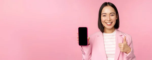 Soddisfatto sorridente donna d'affari asiatica che raccomanda app per telefoni cellulari, società di siti web su smartphone, mostrando schermo e pollici in su, sfondo rosa — Foto Stock