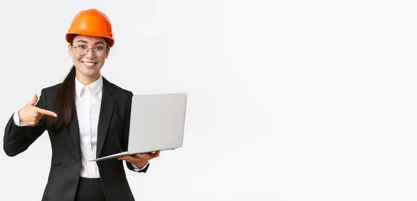 Sonriendo complacido asiático ingeniero jefe mostrando gráfico con beneficio empresarial, haciendo presentación durante la reunión de inversores, usando casco de seguridad, apuntando a la pantalla del ordenador portátil, fondo blanco — Foto de Stock