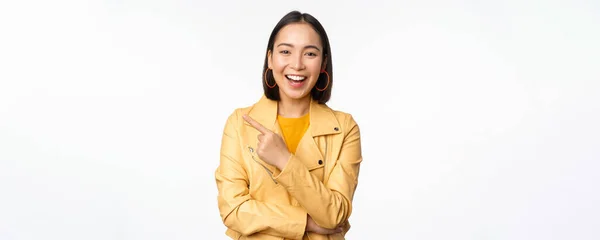 Retrato de hermosa chica coreana riendo, sonriendo y señalando el dedo a la izquierda en el logotipo, mostrando pancarta, de pie en traje casual sobre fondo blanco —  Fotos de Stock