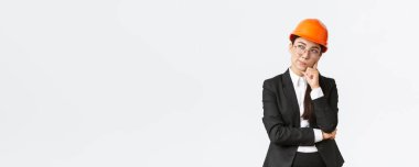 Düşünceli, yaratıcı, Asyalı kadın mimar, inşaat mühendisi, güvenlik kaskı ve takım elbise giyiyor, bina için en iyi seçimi düşünüyor, beyaz arka planda duruyor.