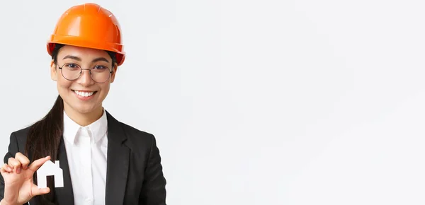 Close-up of happy asian female engingeer, real estate agent in helmet and business suit holding house miniature and smiling, architect working over renovation project, white background — Stock Photo, Image