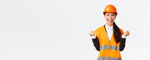 Sikeres győztes ázsiai női építőipari menedzser, mérnök épület látvány visel biztonsági sisak, felemeli a kezét az öröm, ököl szivattyú a teljesítmény és a győzelem, fehér háttér — Stock Fotó