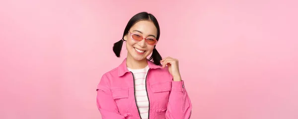 Menina asiática bonita em sunlgasses, sorrindo para a câmera, posando contra o fundo do estúdio rosa — Fotografia de Stock