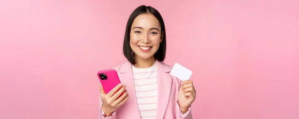 Beeld van glimlachende vrolijke Aziatische zakenvrouw tonen credit card, online betalen op smartphone applicatie, bestellen met mobiele telefoon, staan tegen een roze achtergrond — Stockfoto