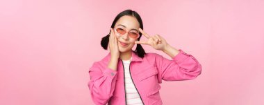 Kawaii Asyalı güneş gözlüklü kızın portresi, barışı gösteriyor, göze yakın V işareti yapıyor ve kameraya sevimli bir şekilde gülümsüyor, pembe arka planda moda kıyafetlerle poz veriyor.