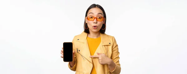Retrato de menina coreana elegante em óculos de sol, sorrindo, apontando o dedo para a tela do smartphone, mostrando aplicativo de telefone móvel, de pé sobre fundo branco — Fotografia de Stock
