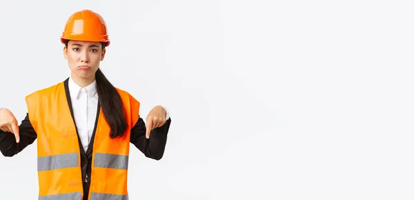 Molesto y sombrío asiático gerente de construcción femenina en casco de seguridad y chaqueta reflectante, señalando con los dedos hacia abajo y pucheros decepcionado, mostrando malas noticias, fondo blanco — Foto de Stock