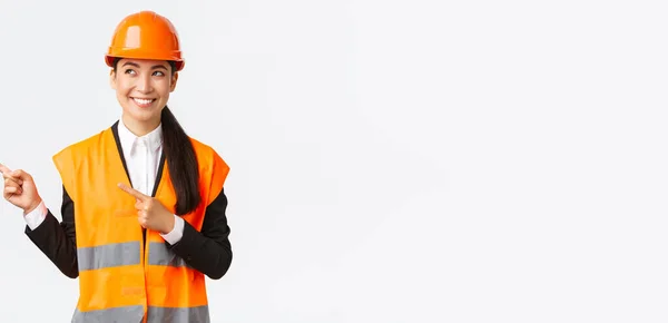 Construção, construção e conceito industrial. Sorrindo confiante asiático engenheiro chefe feminino em capacete de segurança e roupas reflexivas apontando dedos canto superior esquerdo e olhando satisfeito — Fotografia de Stock