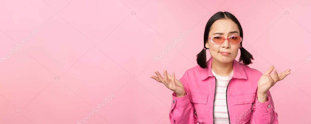 Close up portrait of asian girl looking confused, shrugging puzzled and looking at camera, wearing sunglasses, standing over pink background.