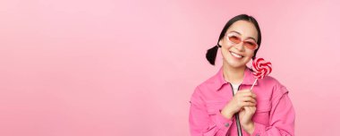 Güneş gözlüklü gülümseyen Asyalı kadın, lolipop şekerleri tutuyor, şeker yiyor ve mutlu görünüyor, pembe arka planda duruyor.