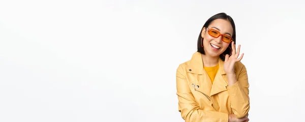 Elegante chica asiática en gafas de sol, sonriendo y buscando fresco, de pie sobre fondo blanco — Foto de Stock