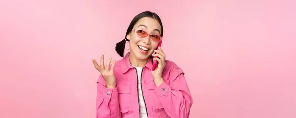 Hermosa modelo femenina coreana en gafas de sol, hablando en el teléfono móvil con la cara feliz, utilizando el servicio celular para llamar a un amigo en el teléfono inteligente, de pie sobre el fondo rosa —  Fotos de Stock