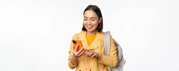 Bild av glad flicka resenär, turist med ryggsäck tittar på smartphone, med hjälp av mobiltelefon rutt app, står över vit bakgrund — Stockfoto