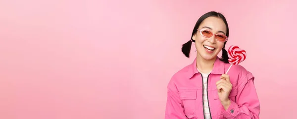 时尚的韩国女孩舔棒棒糖，吃糖果，笑着，站在粉色背景的太阳镜上 — 图库照片