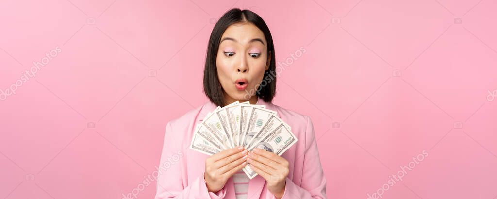 Finance, microcredit and people concept. Happy smiling asian businesswoman showing dollars money, standing in suit against pink background.