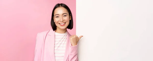 穿着西服的亚裔女商人的画像，女企业经营者用手指指指白色的空墙，贴着信息或广告的木板，站在粉色的背景上 — 图库照片