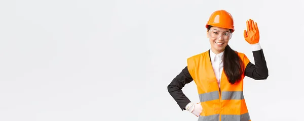 Amistosa mujer asiática sonriente ingeniera de construcción, arquitecta en casco de seguridad, guantes y gafas saludando con la mano, saludando a inversores o clientes en la zona del edificio, fondo blanco — Foto de Stock