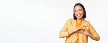 Asyalı güzel bir kızın görüntüsü kalp, aşk işareti, özen ve romantik duygular gösteriyor, beyaz stüdyo arka planında duruyor.