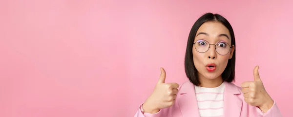 Primer plano retrato de mujer corporativa impresionada, mujer de negocios asiática en gafas, mostrando los pulgares hacia arriba, mirando sorprendido a la cámara, recomendando, fondo rosa —  Fotos de Stock