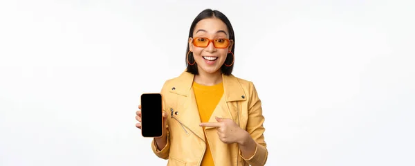 Retrato de menina coreana elegante em óculos de sol, sorrindo, apontando o dedo para a tela do smartphone, mostrando aplicativo de telefone móvel, de pé sobre fundo branco — Fotografia de Stock