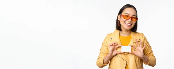 Elegante chica asiática atractiva en gafas de sol, mostrando la tarjeta de crédito y sonriendo, de pie feliz contra fondo blanco estudio — Foto de Stock