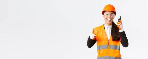 Soddisfatto felice sorridente asiatica ingegnere femminile, tecnico industriale in casco di sicurezza e uniforme mostrando pollici in su mentre loda grande lavoro utilizzando walkie-talkie, dare il permesso di lavorare — Foto Stock