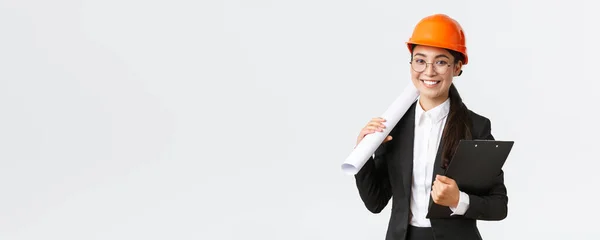 Exitosa arquitecta asiática en traje de negocios y casco sosteniendo planos y portapapeles con notas, inspector mirando obras de construcción, sonriendo a la cámara, fondo blanco — Foto de Stock