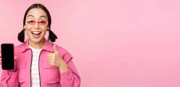 Feliz chica con estilo recomienda la aplicación en el teléfono móvil. Sonriendo modelo femenino asiático mostrando la pantalla del teléfono inteligente y el pulgar hacia arriba, de pie sobre el fondo rosa —  Fotos de Stock