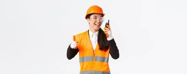 满意的笑容满面的亚洲女工程师，戴着安全帽、身穿制服的工业技术人员，在称赞使用对讲机所做的出色工作的同时，允许工作 — 图库照片