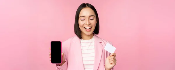Mulher de negócios coreana sorridente em terno, mostrando tela do telefone móvel, cartão de crédito, mostrando interface de aplicativos bancários on-line, fundo rosa — Fotografia de Stock