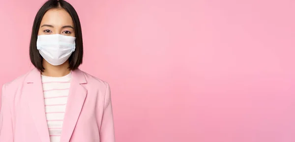 Porträtt av asiatisk affärskvinna i medicinsk ansiktsmask, bära kostym, begreppet kontorsarbete under covid-19 pandemi, stående över rosa bakgrund — Stockfoto