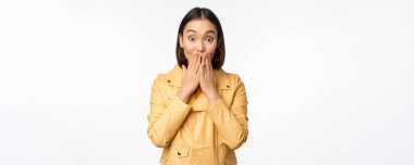 Kameraya ilgi duyan heyecanlı Asyalı bir kızın portresi, büyük haberlere ya da duyuruya hayret verici bir tepki, beyaz arka planda sarı ceketin içinde duruyor.